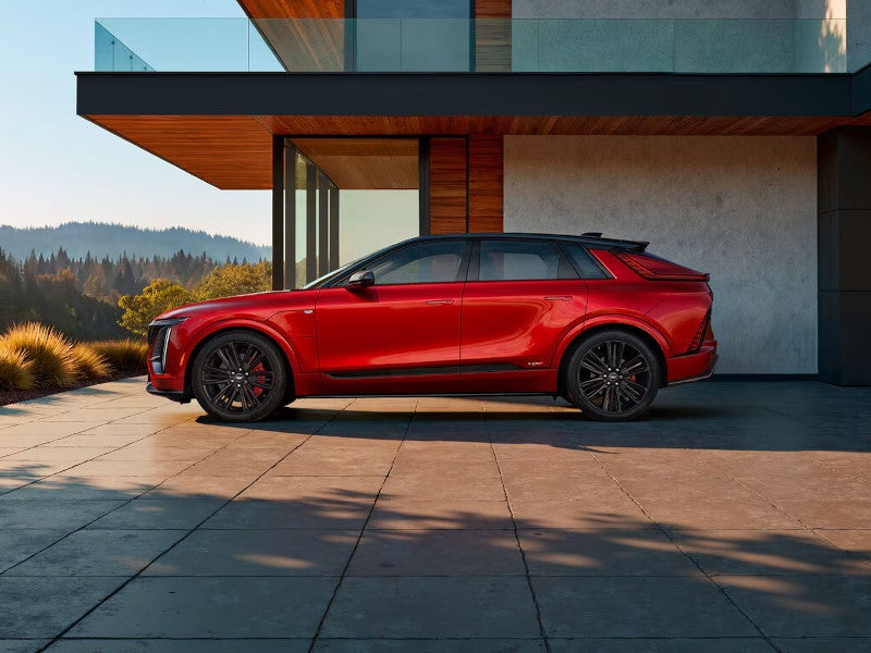 Side profile of a red Cadillac Lyriq electric SUV parked in front of a modern home with forested hills behind it.